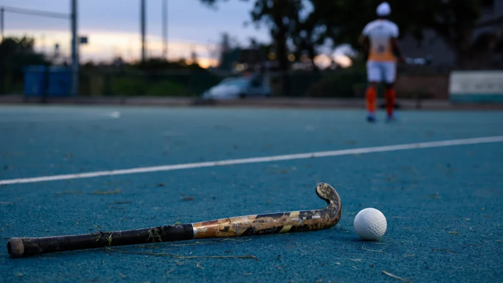 A field hockey stick and ball on a blue turf with a player in the background, illustrative image.