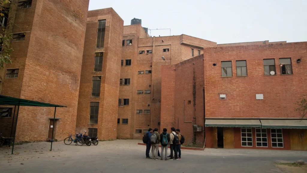 Red-brick JNU academic buildings with students in a campus courtyard. AI-generated image grounded in a real Wikimedia Commons photo of JNU campus buildings.
