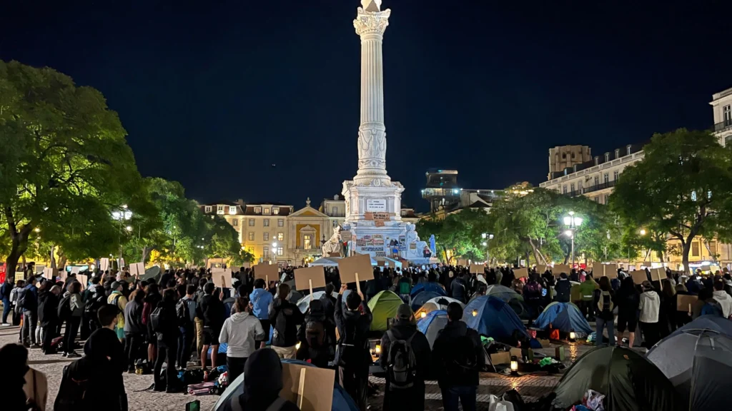 Housing protest camp in Rossio square, Lisbon, at night. AI-generated image grounded in a real 2026 protest photo (Afonso Dimas Martins, Wikimedia Commons, CC BY-SA 4.0).