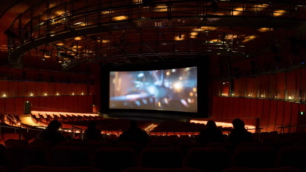 A grand, empty cinema auditorium lit in warm red and gold tones ahead of a mythological epic premiere, with dramatic light falling across the seats and screen.