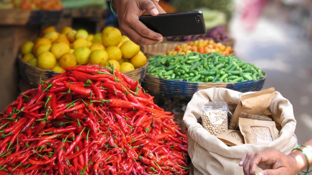 Hands holding a smartphone over vegetable baskets at an Indian market.