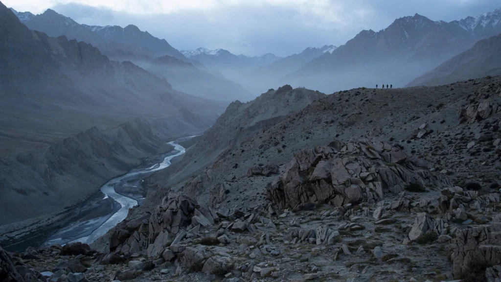 A rugged Himalayan mountain valley at dawn, with distant figures on a ridge.