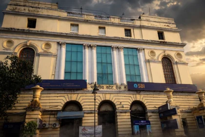State Bank of India branch building with blue SBI signage. Photo: Pinakpani / Wikimedia Commons, CC BY-SA 4.0 (editorial background composite).