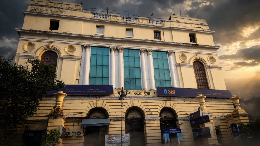 State Bank of India branch building with blue SBI signage. Photo: Pinakpani / Wikimedia Commons, CC BY-SA 4.0 (editorial background composite).
