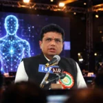 D. Sridhar Babu at a technology conference stage with a digital avatar on screen. Composite: real photo of the minister (Wikimedia Commons) with AI-generated conference background.