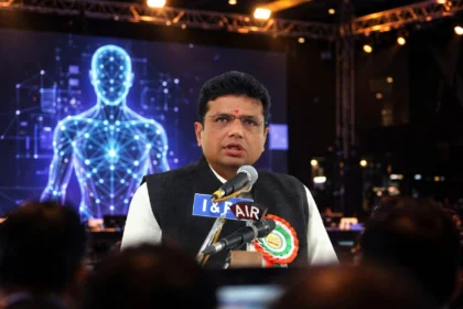 D. Sridhar Babu at a technology conference stage with a digital avatar on screen. Composite: real photo of the minister (Wikimedia Commons) with AI-generated conference background.