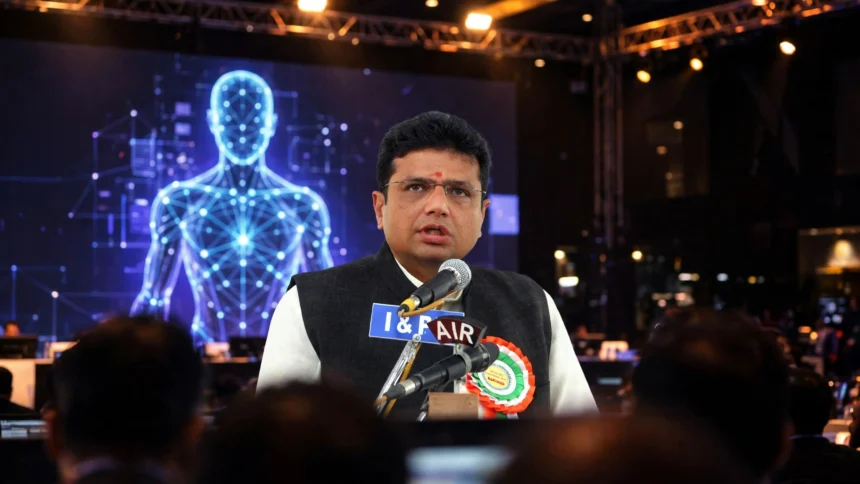 D. Sridhar Babu at a technology conference stage with a digital avatar on screen. Composite: real photo of the minister (Wikimedia Commons) with AI-generated conference background.
