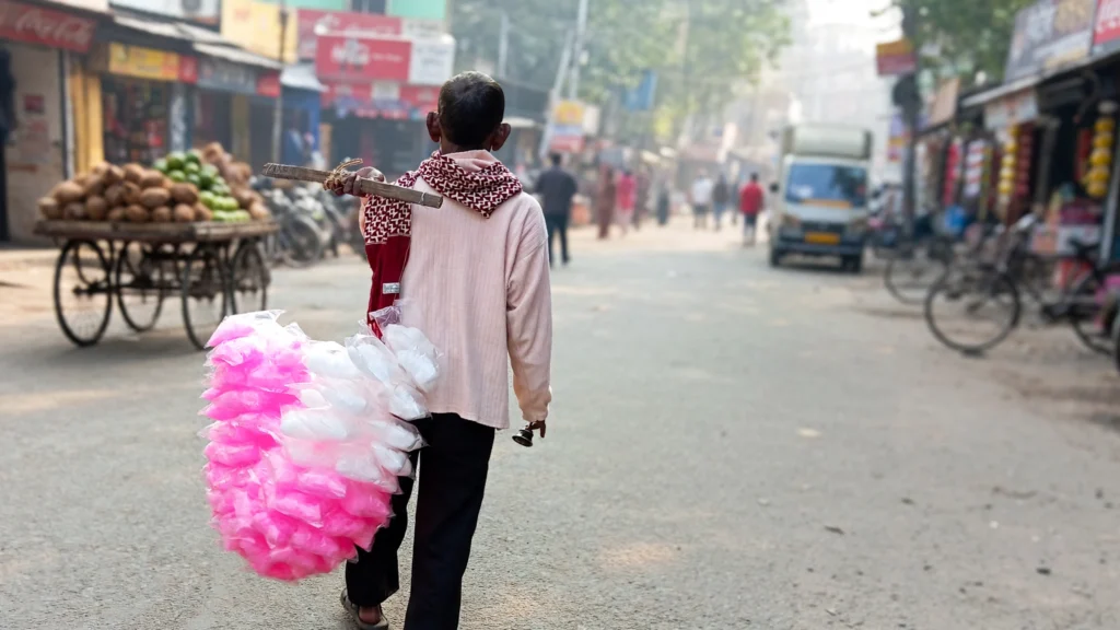 Cotton candy seller carrying pink and white cotton candy packs on a city street. AI image grounded in a real Wikimedia Commons photo of a cotton candy seller in Kolkata.
