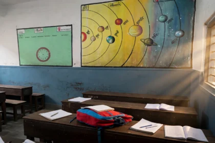 Indian school classroom in warm morning light, desks with notebooks and school bags, planets wall chart. Classroom reference photo: Anindya202414 / Wikimedia Commons (CC BY-SA 4.0), background composite