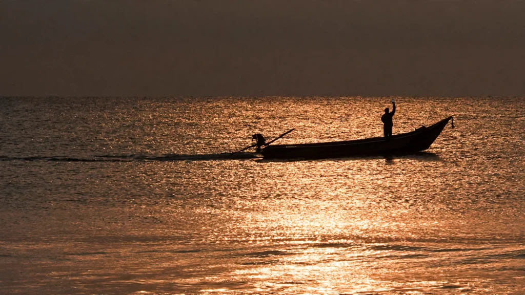 A fisherman holds up a mobile phone from a boat at sunrise off Chennai, illustrating satellite messaging for areas beyond mobile coverage. Grounded in a real photo by Tagooty / Wikimedia Commons.